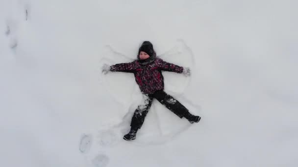 Vista superior de niño niño acostado en el suelo de nieve y hace figura de ángel, divertido concepto de la infancia — Vídeos de Stock