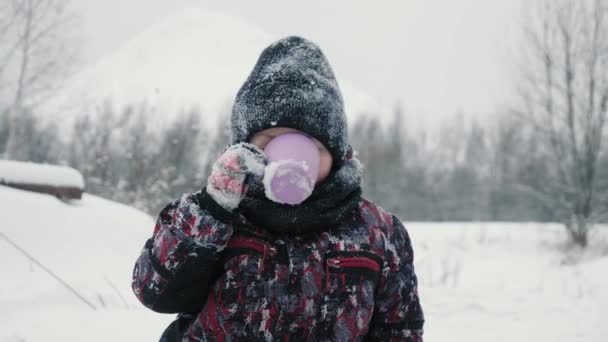 Χαρούμενο αγόρι πίνοντας ζεστό τσάι από το κύπελλο στο χιονισμένο δάσος κατά τη χειμερινή βόλτα. Αγόρι απολαμβάνει το χειμερινό τσάι, ενώ το περπάτημα στο κρύο ημέρα στο δάσος το χειμώνα. Αργή κίνηση — Αρχείο Βίντεο