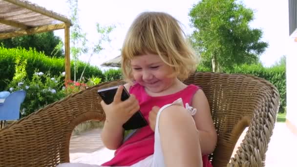Ребенок смотрит телефон, сидя в плетеном кресле — стоковое видео