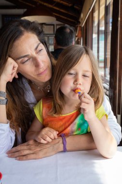 Dört yaşında, sarışın küçük bir kız. Lolipop yiyor. Lolipopun üstünde oturuyor.
