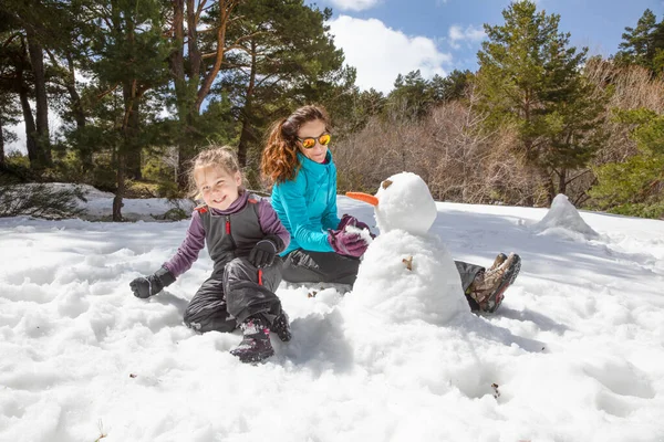 快乐的家庭4岁的金发姑娘和女人坐在雪地上笑着 在森林里做了个雪人 鼻子是胡萝卜 — 图库照片