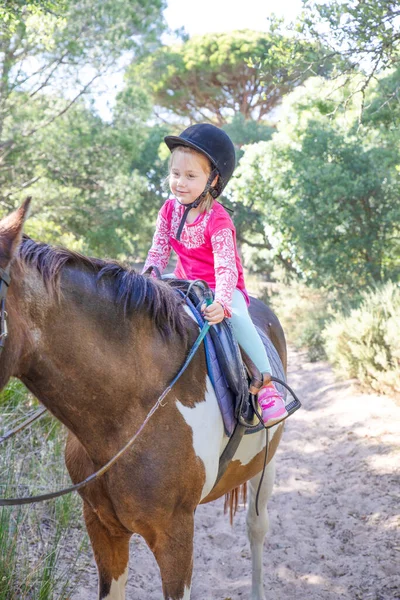 little girl four years old with pink shirt and black cap riding catching the reins of the horse in a forest