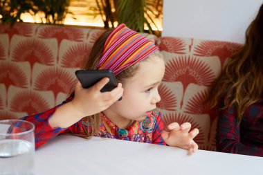 Yedi yaşında, saç bandı takan, kırmızı koltukta oturan, kulaklarının yanındaki akıllı telefonu dinleyen tatlı bir kız.