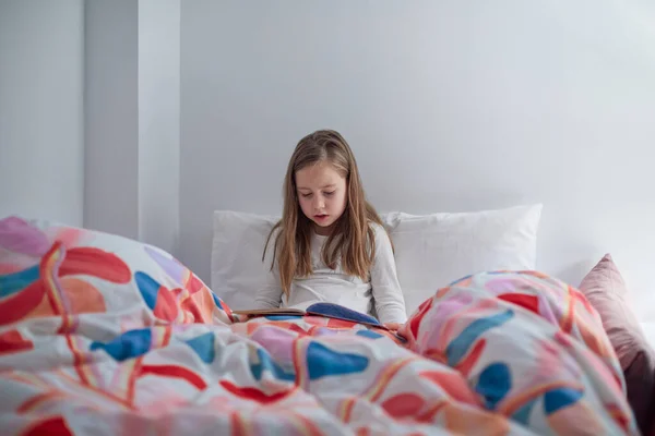 7岁的可爱的金发女孩躺在床上看书或看漫画 带着枕头和大大的羽绒被 — 图库照片