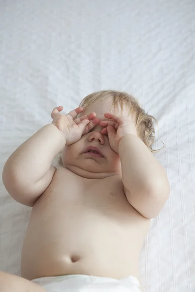 Baby touching eyes on white bed — Stockfoto