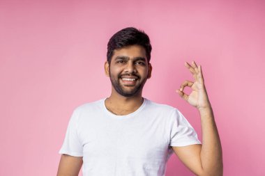 Vücut dili kavramı. Beyaz t-shirt giyen neşeli Hintli erkek öğrenci, tamam işareti yapıyor, sınav sonuçlarından tatmin oluyor, neşeyle gülümsüyor, pembe arka planda soyutlanıyor..