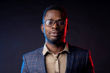 Gözlüklü, gömlekli, takım elbiseli, siyah arkaplana karşı duran, kameraya bakan, kendine güveni tam, ciddi sakallı Afrikalı iş adamının yakın portresi..