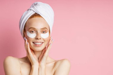 Göz şişkinliğini ve koyu halkaları azaltmak için yamalar yapıştıran, her iki avuçla nazikçe yüzyüze dokunan, pembe arka planda çıplak poz veren şehvetli güzel bir kadının vesikalığı. Güzellik konsepti.