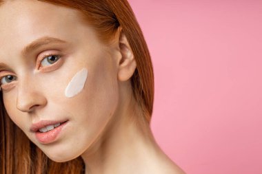 Yanağında krema olan genç bir kadının portresi. Kızıl saçlı, taze çilli, pembe duvarın karşısında mutlu bir şekilde gülümseyen beyaz bir kız. Doğal güzellik konsepti.