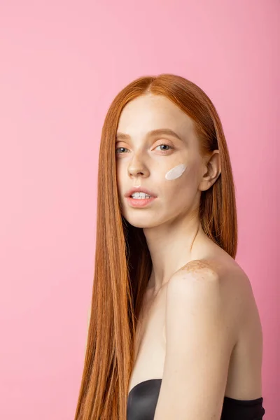 Stock image Vertical shot of gorgeous sensual caucasian woman with perfect soft skin, shiny long red hair, touching neck, posing on pink background. Beauty model with cream or mask on cheek.