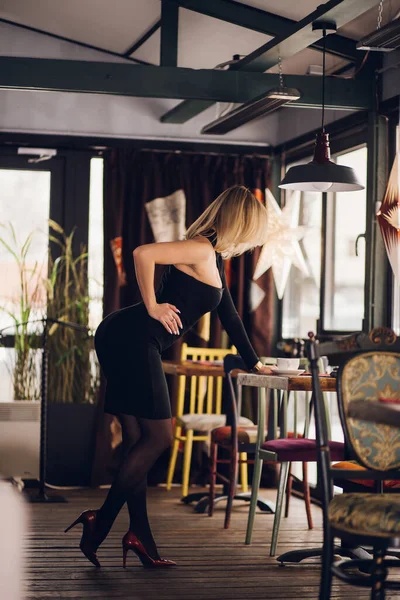ブロンドの女性が横に レストラン カフェのテーブルの上にもたれ立って 短い黒いドレスを着て 赤い靴 美しい姿のセクシーな女の子 全長写真 — ストック写真