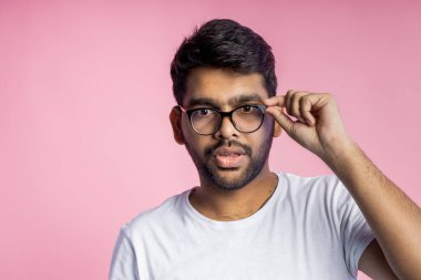 Kötü görüşlü Hintli genç bir adam, gözlükleriyle iyice bakıyor, bir şeyler okumaya çalışıyor, göz çerçevesine dokunuyor, günlük kıyafetler giyiyor, pembe arka planda fotokopi alanı ile izole edilmiş..