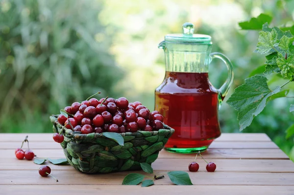 Cold refreshing drink from cherries in a pitcher and ripe berries in basket on wooden table in the garden — Stock Photo, Image