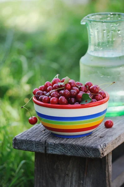Jug of cold fresh water with cherries on wooden table in the garden — Stock Photo, Image