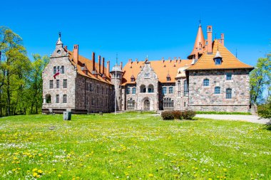Cesvaine, Letonya - 05.23.2020: Cesvaine kalesi - 19. yüzyılın sonlarında bir malikane, kahverengi kiremit çatılı taşlardan yapılmış bir bina, yeşil çimenli bir çimen ve sarı karahindibalar..
