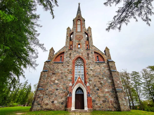 Cesvaine, Letonya - 07.23.2019: Cesvaine Lutheran Kilisesi, yeşil ağaçlarla çevrili mavi gökyüzüne karşı, saatli renkli taşlardan oluşan bir bina.