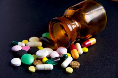 Kapsül şeklinde ilaçlar ve haplar yan tarafına düşmüş siyah cam bir kavanozun yanında yatıyor..
