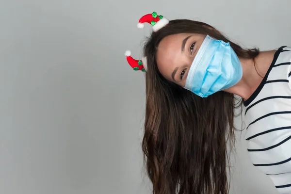Una giovane donna con una maschera medica sbircia fuori da dietro la cornice. Natale durante la pandemia di coronavirus. — Foto Stock