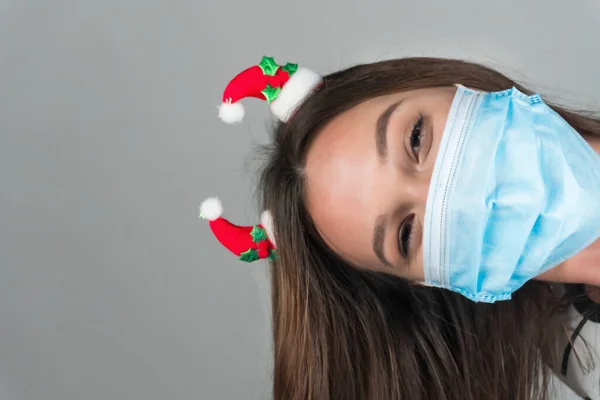 Una giovane donna con una maschera medica sbircia fuori da dietro la cornice. Natale durante la pandemia di coronavirus. — Foto Stock