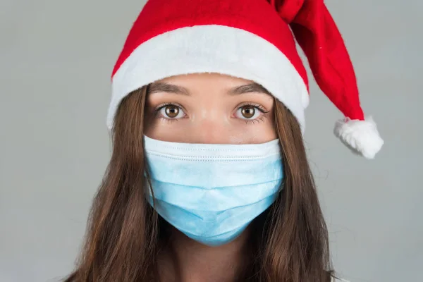 一个戴着圣诞老人帽和医疗面具的年轻女子看着摄像机。在大流行病中庆祝新年. — 图库照片
