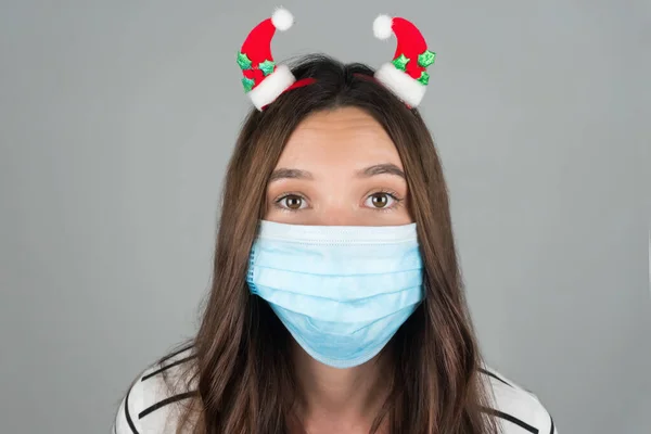 Una giovane donna con una maschera medica con una decorazione natalizia in testa. Fondo grigio — Foto Stock