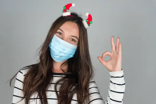 Giovane donna in maschera medica per Natale mostra gesto ok. Festeggiare il Natale nel bel mezzo di una pandemia. — Foto Stock