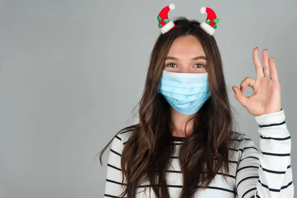 Giovane donna in maschera medica per Natale mostra gesto ok. Festeggiare il Natale nel bel mezzo di una pandemia. — Foto Stock