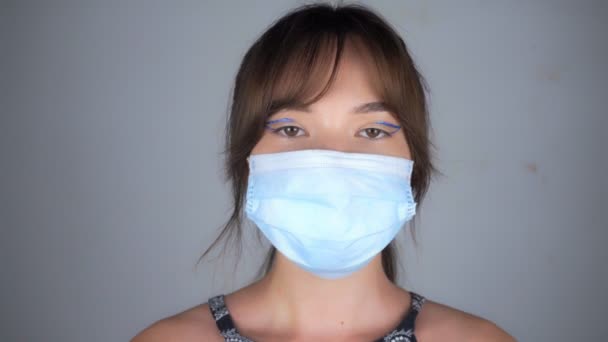 Νέα όμορφη γυναίκα με μακιγιάζ και σε μια ιατρική μάσκα κοιτάζει την κάμερα — Αρχείο Βίντεο
