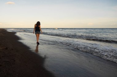 Genç bir kadın gün batımında deniz kıyısında yürüyor..