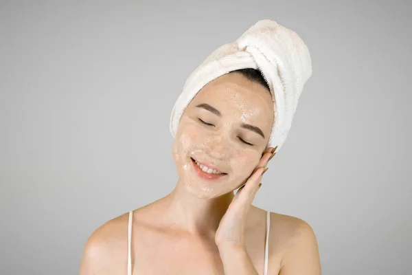 Ευτυχισμένη νεαρή γυναίκα με πετσέτα μπάνιου στο κεφάλι απλώνει αφρό καθαρισμού. — Φωτογραφία Αρχείου