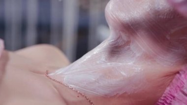 Kadın güzellik uzmanı elinde fırçayla genç bir kadının yüzüne maske takıyor..