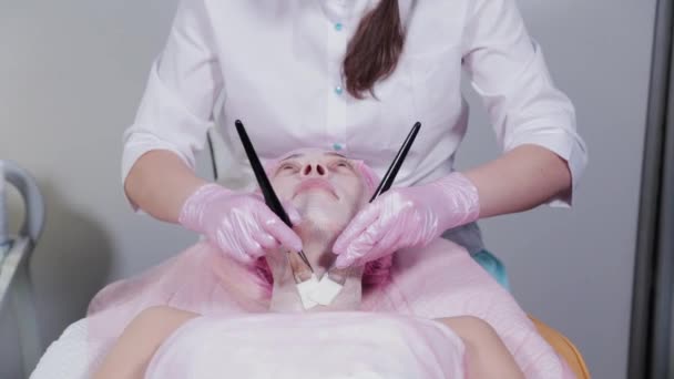 Женщина-косметолог наносит маску на лицо молодой женщины кистью. — стоковое видео