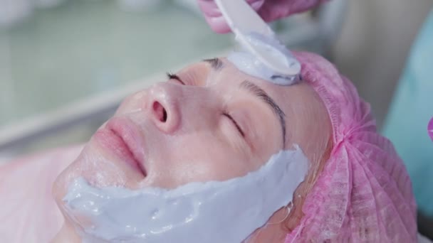 Женщина-косметолог наносит маску на лицо молодой женщины со специальной лопаткой. — стоковое видео