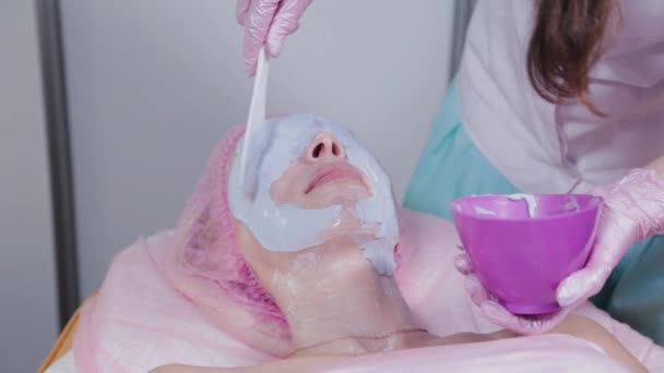 Женщина-косметолог наносит маску на лицо молодой женщины со специальной лопаткой. — стоковое видео