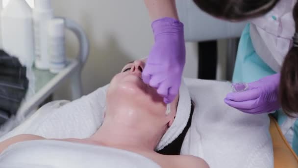 Женщина-косметолог применяет пилинг к женщине лицом к клиенту косметологической клиники. — стоковое видео
