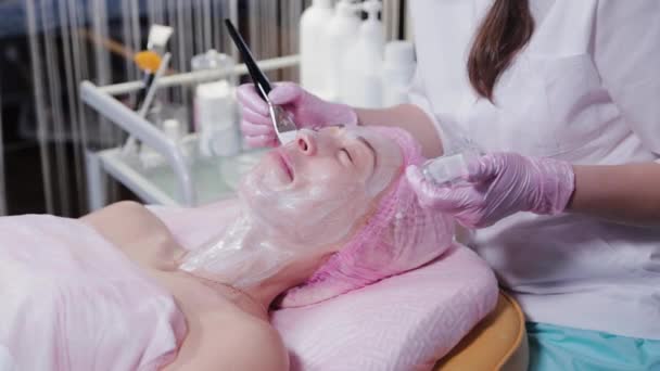 Женщина-косметолог наносит маску на лицо молодой женщины кистью. — стоковое видео