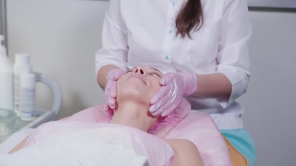 Schoonheidsspecialiste smeert crème op het gezicht van een vrouwelijke patiënt. Gezichtsmassage. — Stockvideo