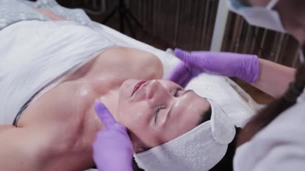 Femme esthéticienne frotte crème sur le visage d'un client d'une clinique de cosmétologie. — Video