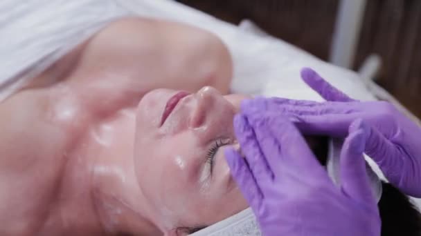 Femme esthéticienne frotte crème sur le visage d'un client d'une clinique de cosmétologie. — Video