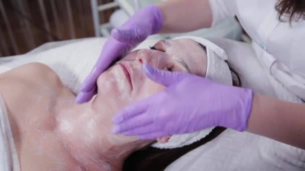 Žena kosmetička rozmazává krém na tváři pacientky. Masáž obličeje. — Stock video