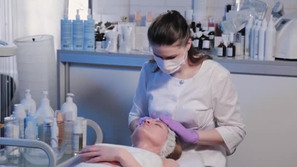 Kvinna kosmetolog gnider grädde på kvinnor ansikte. — Stockvideo