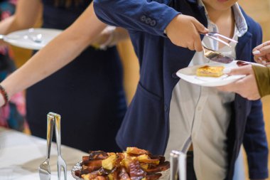 Mavi ceketli ve beyaz gömlekli genç, kapalı alandaki bir kutlama etkinliğinde açık büfe masasından kendine bir hamur işi servis ediyor.