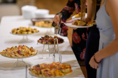 İnsanlar, özel bir etkinlik sırasında uzun bir masanın üzerine serilmiş çeşitli lezzetli aperatifler ve lezzetli tartlardan oluşan bir büfeden kendilerine servis yapıyorlar ya da kapalı bir yerde yumuşak ortam aydınlatmasıyla kutluyorlar.