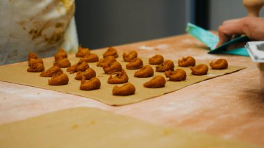 Profesyonel bir mutfak ortamında pişirmeden önce ahşap masadaki parşömen kağıdına yerleştirilmiş küçük pişmemiş hamur topları hazırlayan eller.
