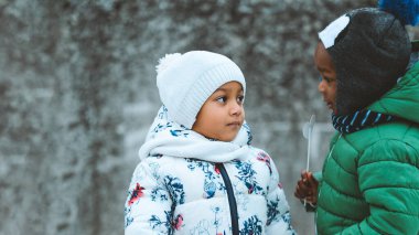 Sıcak kış giysileri giymiş iki genç, soğuk bir kış gününde karda konuşarak havanın tadını çıkarıyorlar.