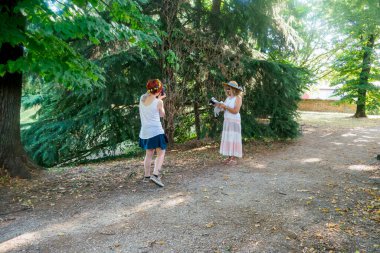 Parkta güneşli güzel bir günün tadını çıkaran iki kadın yemyeşil ağaçlar ve yeşilliklerle çevrili doğada bir kamera ve telefonla anı yakalıyorlar..