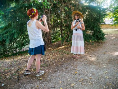 İki kadın açık havada güneşli bir günün tadını çıkarırken, biri elinde ağaçlar ve yapraklarla çevrili küçük beyaz bir kedi yavrusu tutarak büyüleyici ve doğal bir manzara yaratıyor.