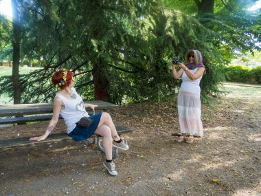 Parkta güneşli bir günün tadını çıkaran iki genç kadın, tahta bir bankta dinlenirken Retro kamerayla anıları yakalıyorlar.