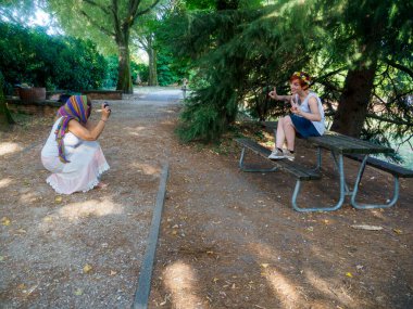 Parkta piknik masası ve ağaçlarla fotoğraf çeken iki kadın güneşli bir günde doğal ve huzurlu bir ortam yaratıyor.