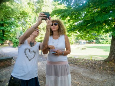 Güneşli bir öğleden sonra, yeşil ağaçlar ve benekli güneş ışığıyla birlikte parkta selfie çeken iki kadın, tatil fotoğrafçılığı için sıcak ve davetkar bir ortam yaratıyor.